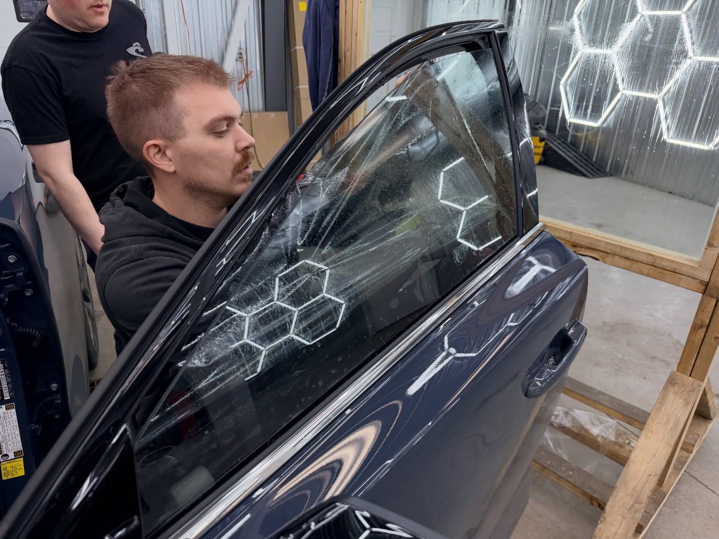 Person opening a car door in a workshop, with a cracked window and hexagonal light reflections.