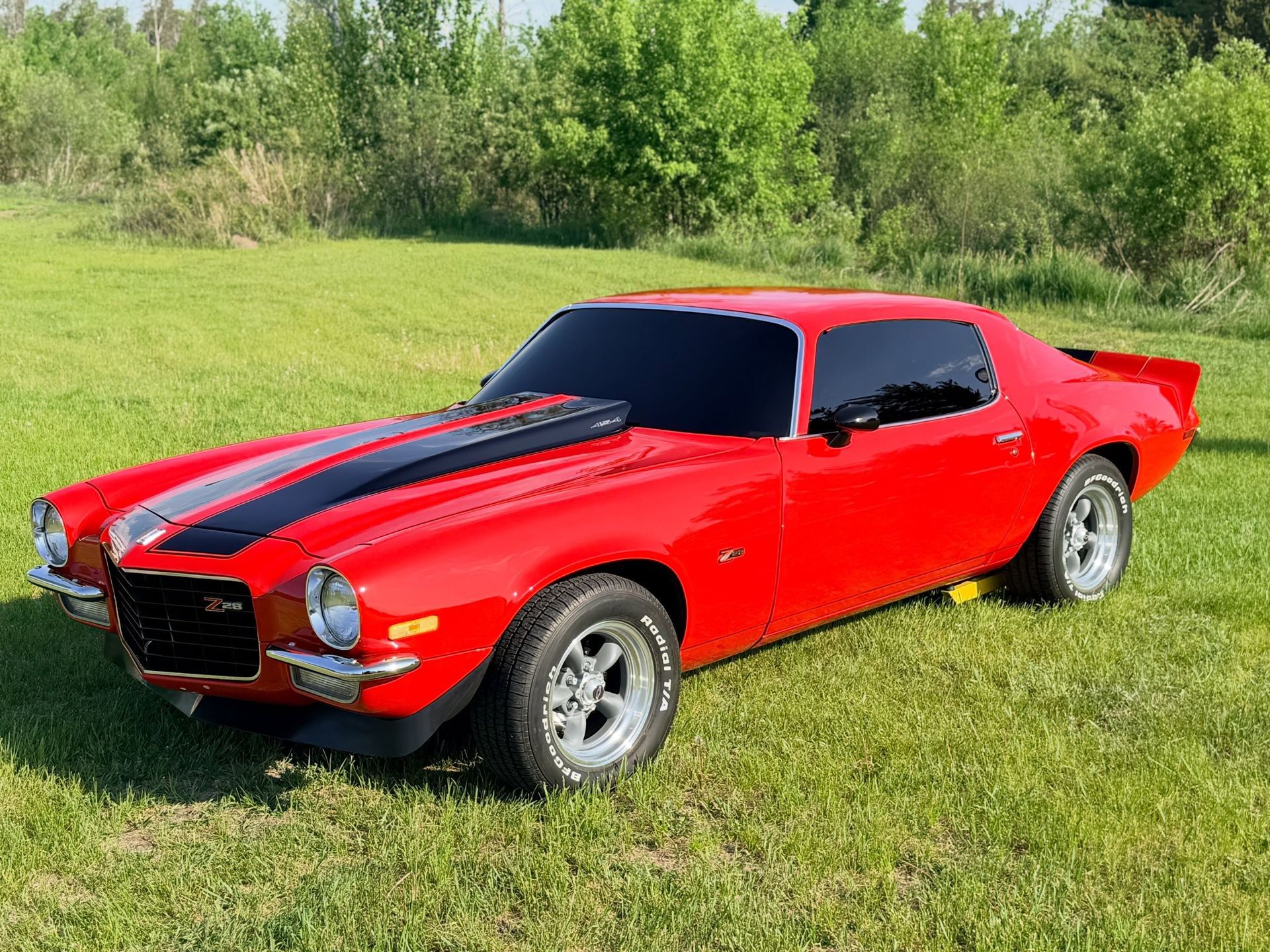Red classic muscle car with black racing stripes parked on grass near trees
