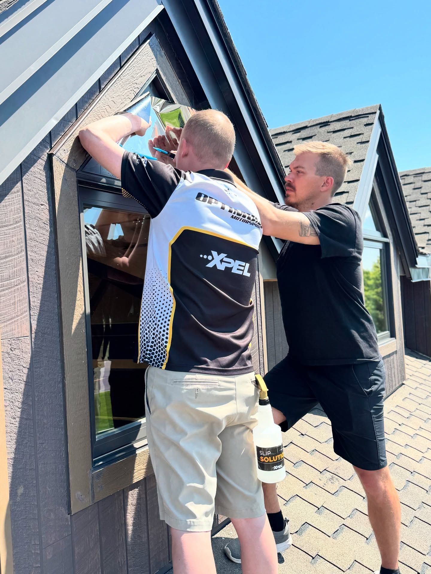 Two workers installing a black-framed skylight on a roof, one inside the opening and one holding the frame.