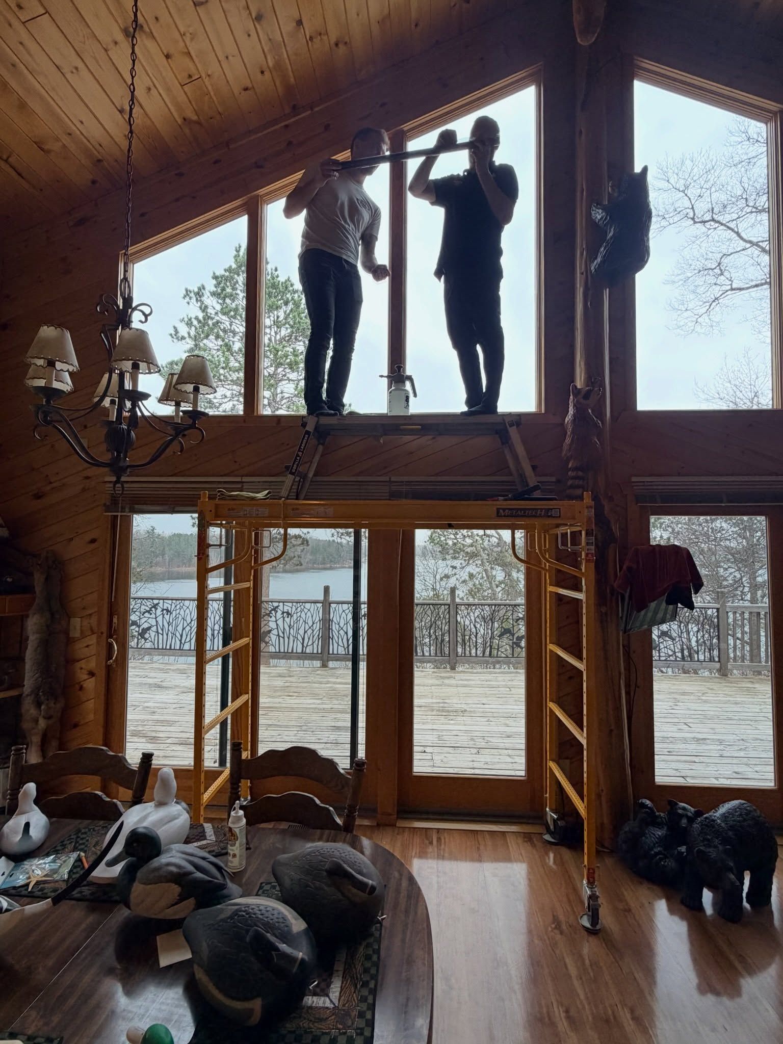 Two people work on a window inside a rustic cabin with a snowy lake view outside.