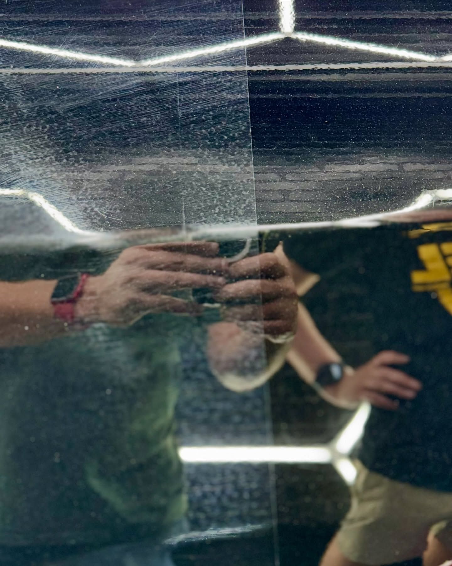 Mirrored reflection of two people in a gym, one holding a phone, with bright overhead lights.