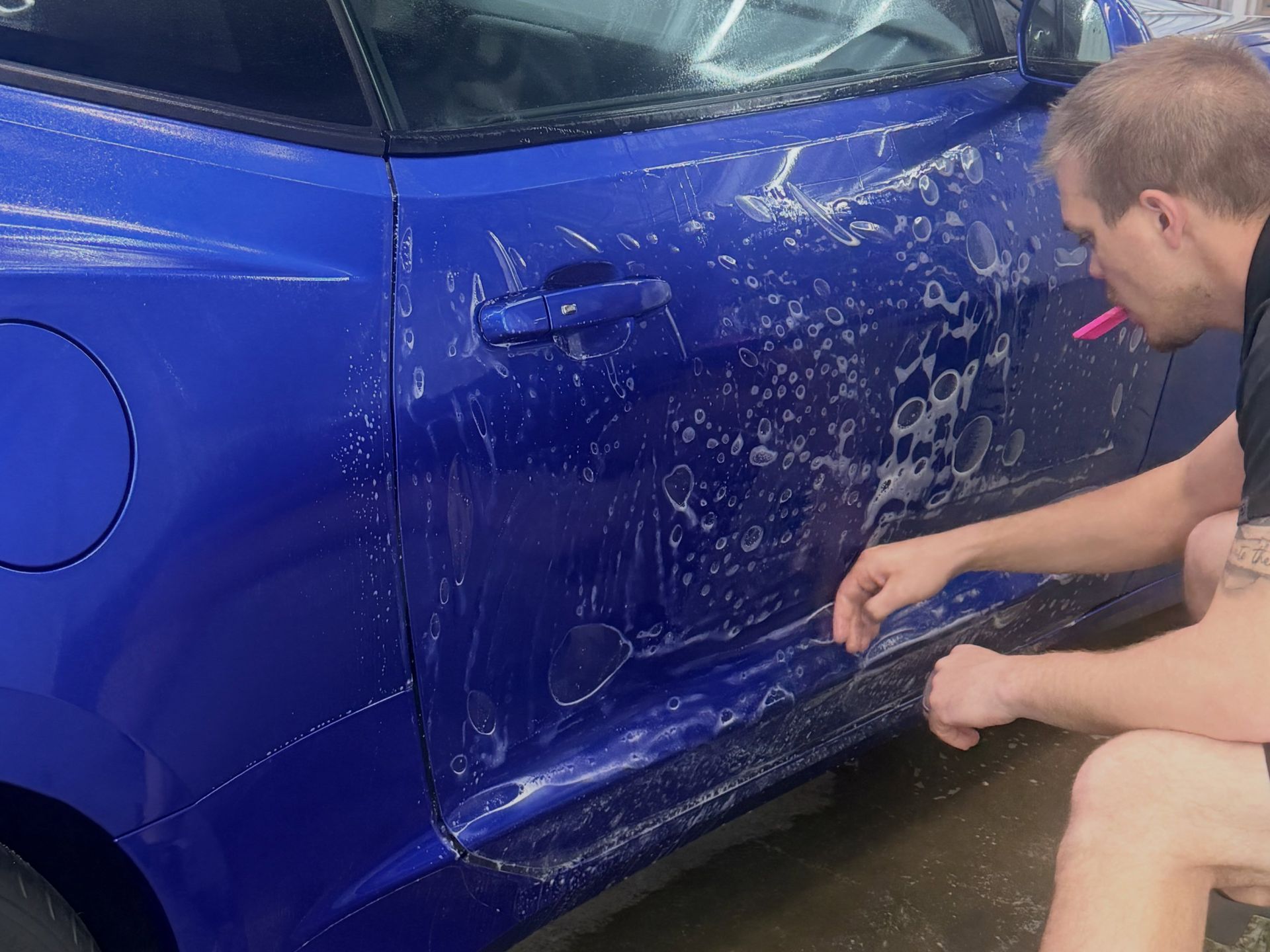 Man scrubs soap off a blue car door in a driveway, with suds and water on the panel.