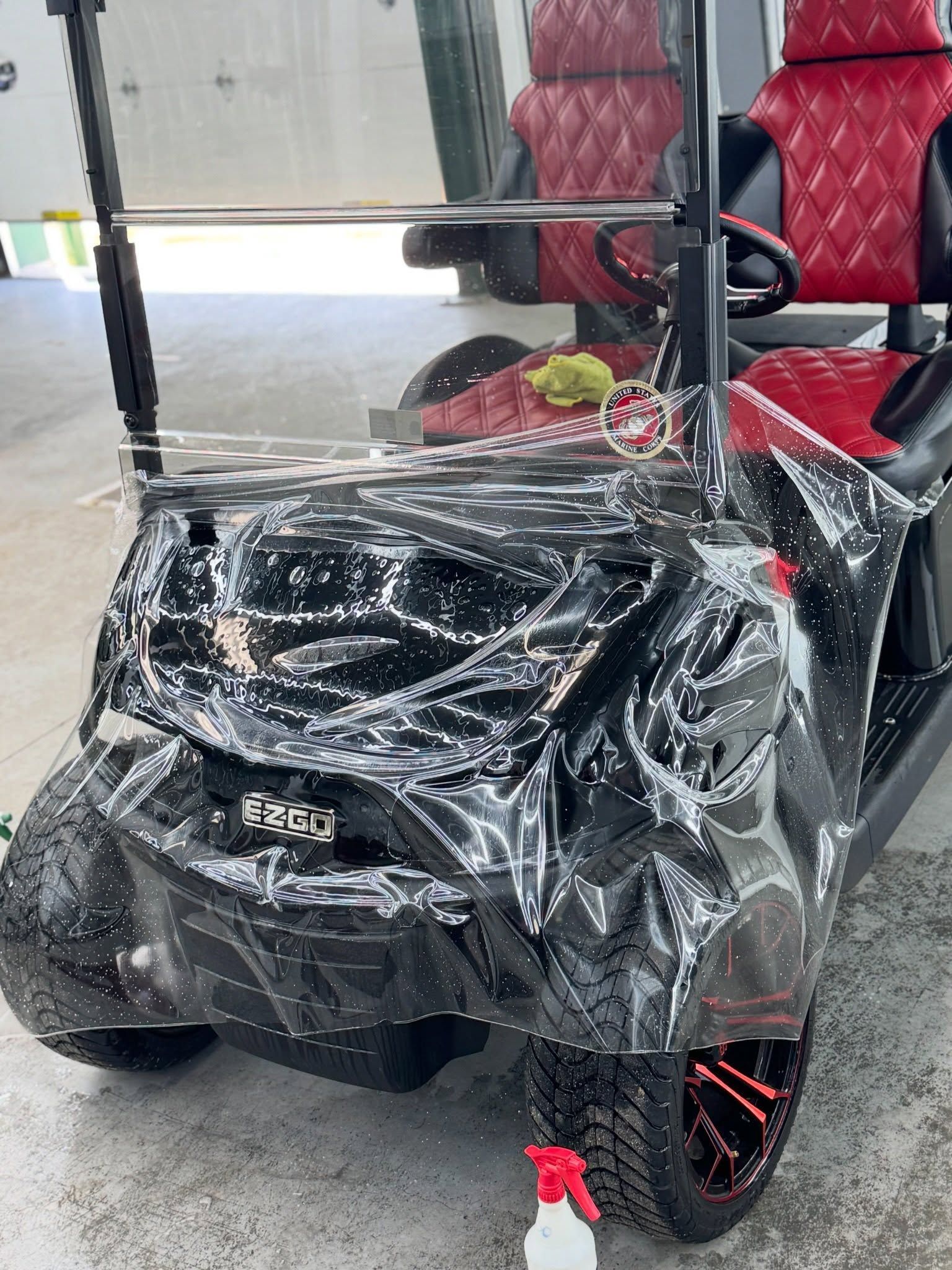 Black golf cart wrapped in clear plastic, with red seats and orange-rimmed wheels, indoors.