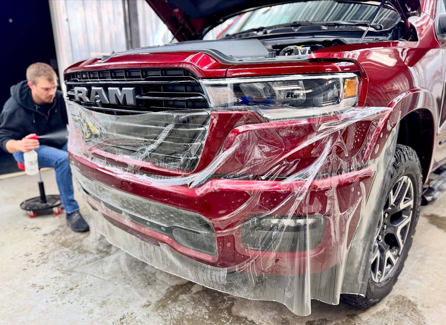 Red RAM truck front wrapped in protective film while a person crouches beside it in a workshop