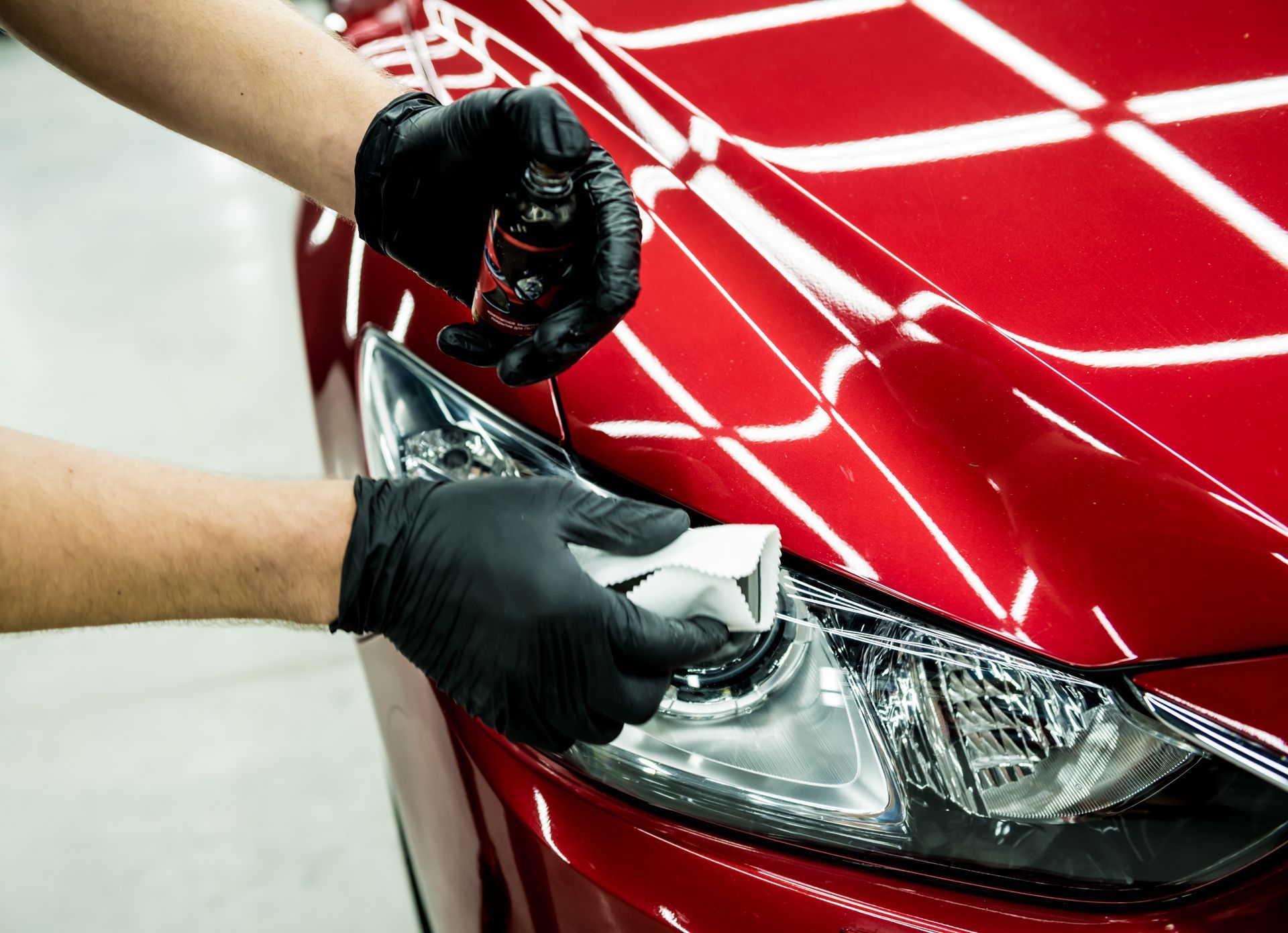Gloved hands polishing a red car’s headlight and hood with a cloth