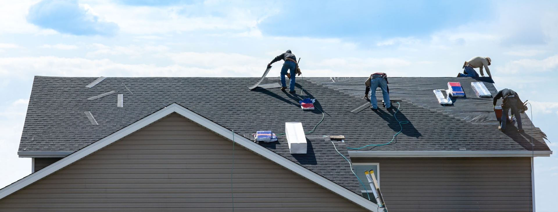 Workers on a shingled roof using tools and equipment, with shingles stacked nearby under a cloudy sky