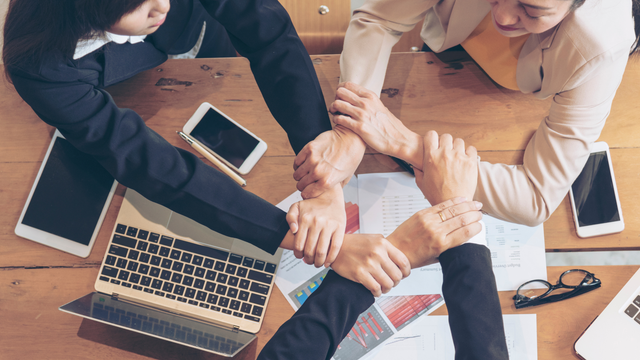 Hands clasped in unity around a table with phones, a laptop, and papers.