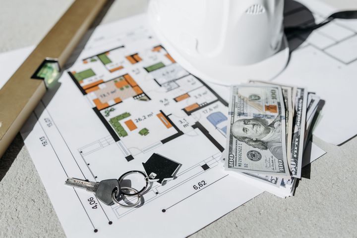 House keys, a stack of dollar bills, and a hard hat on a house floor plan