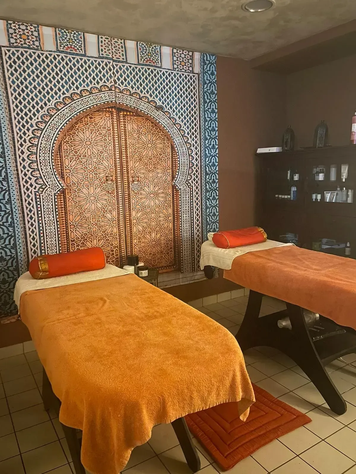 Sala massaggi con due lettini ricoperti di tessuto arancione, arazzo a parete decorato e scaffali con prodotti per la spa.