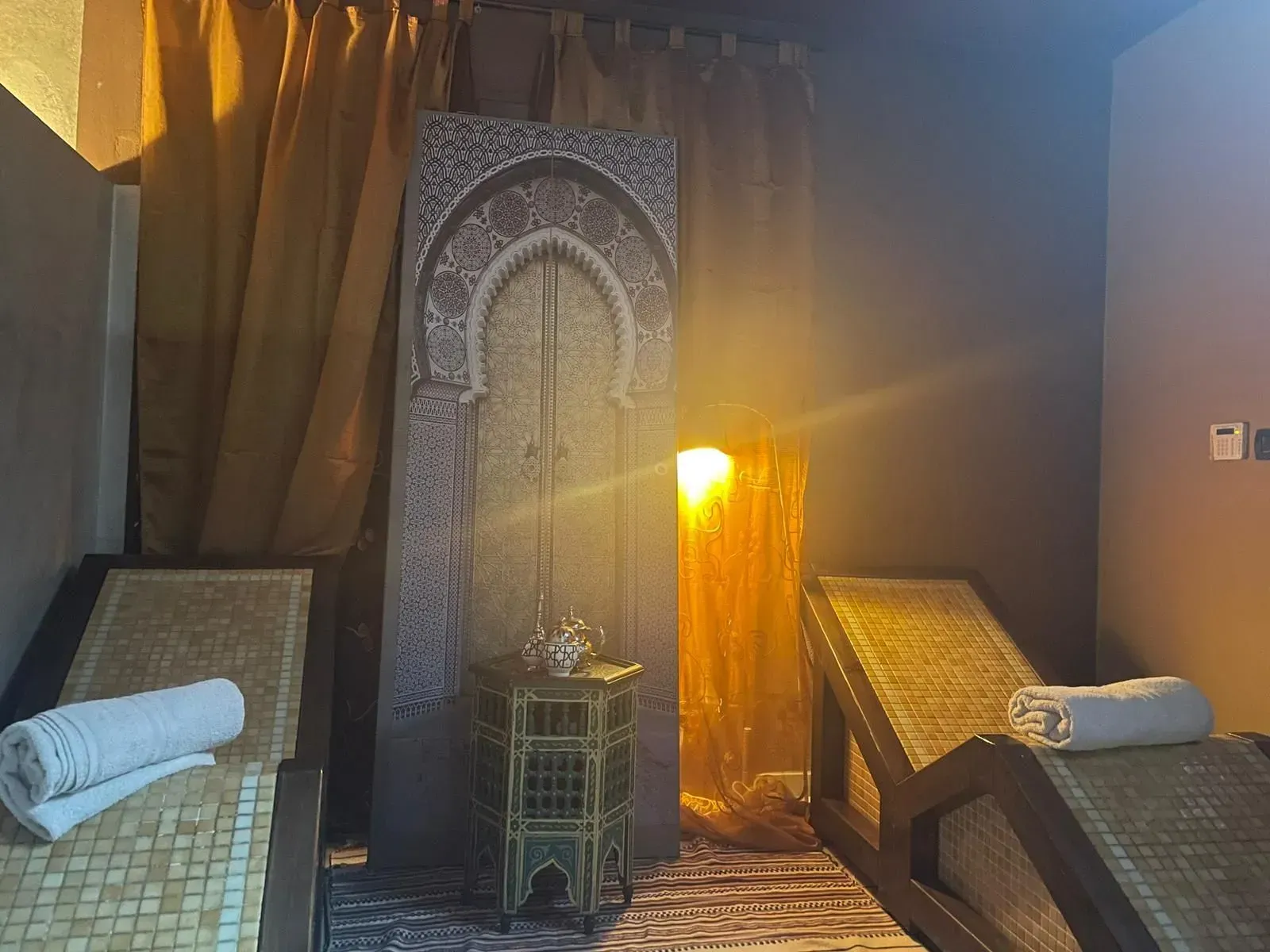Una stanza accogliente con due lettini prendisole, una lampada soffusa e un pannello decorativo ad arco tra le tende.