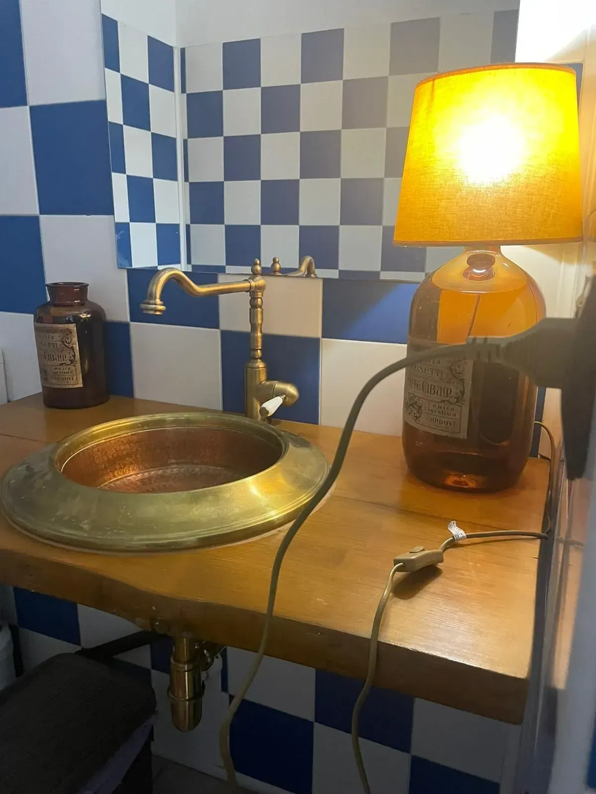Lavabo del bagno con rubinetto in ottone, lampada a bottiglia color ambra e parete rivestita di piastrelle blu e bianche.