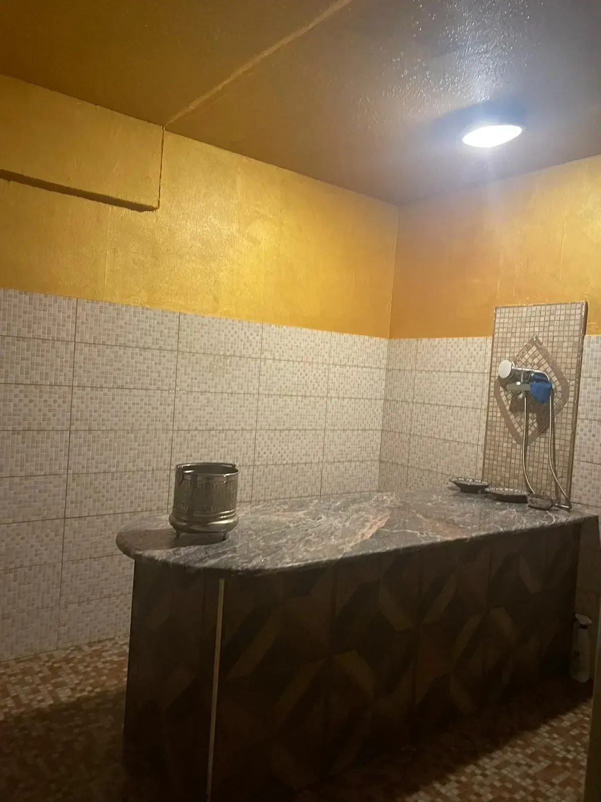 Piccola stanza interna con pareti rivestite di piastrelle gialle e bianche, un bancone e un lavabo a parete sotto una lampada.