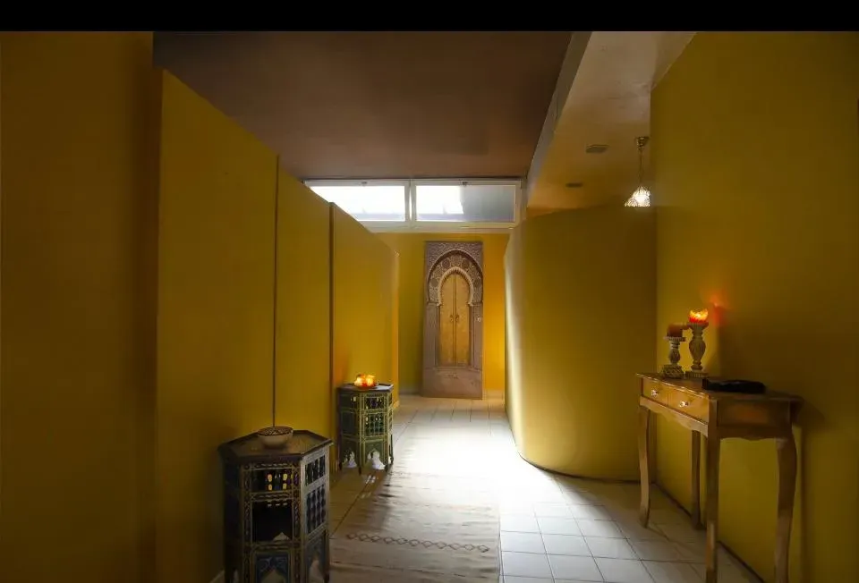 Stretto corridoio giallo con porte di legno, tavolini e una candela accesa sulla destra.