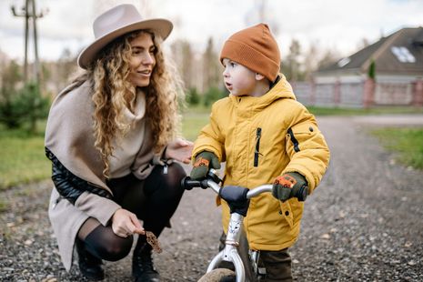 A woman is kneeling down next to a little boy riding a bike.