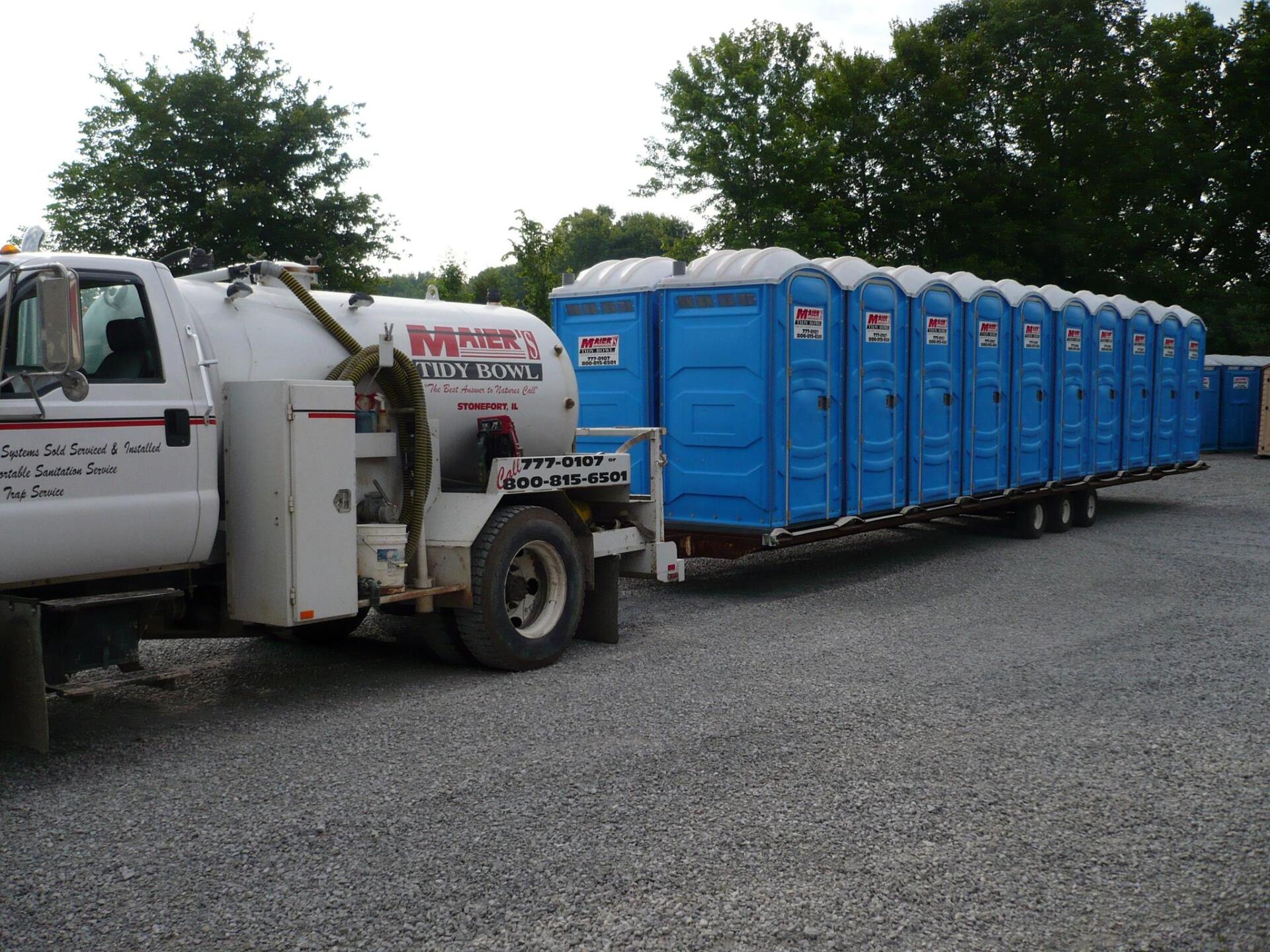 White truck hauling a long row of blue portable toilets in a gravel lot