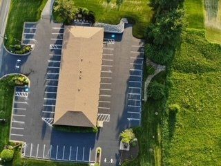 Aerial view of a long, brown-roofed building with ample parking, adjacent to a green field.