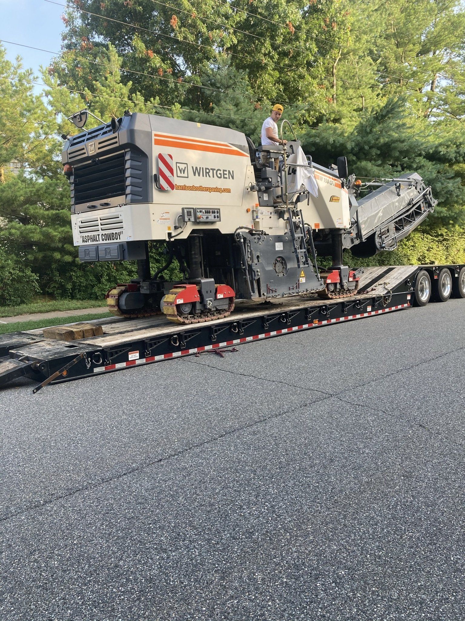 Road milling machine on a flatbed trailer, operator on the machine, parked on asphalt surface.