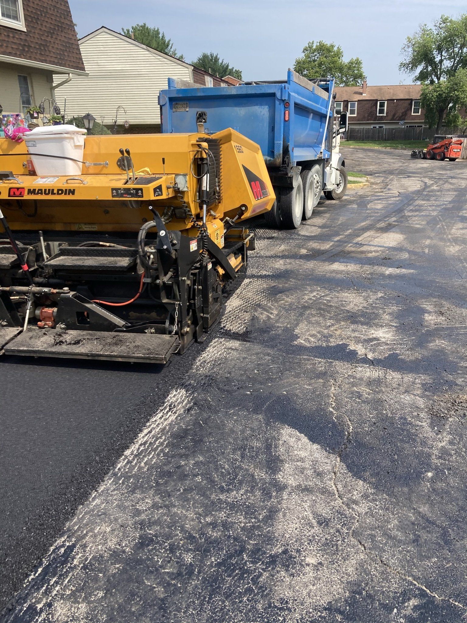 Asphalt paving machine laying blacktop on a street, with a dump truck nearby.