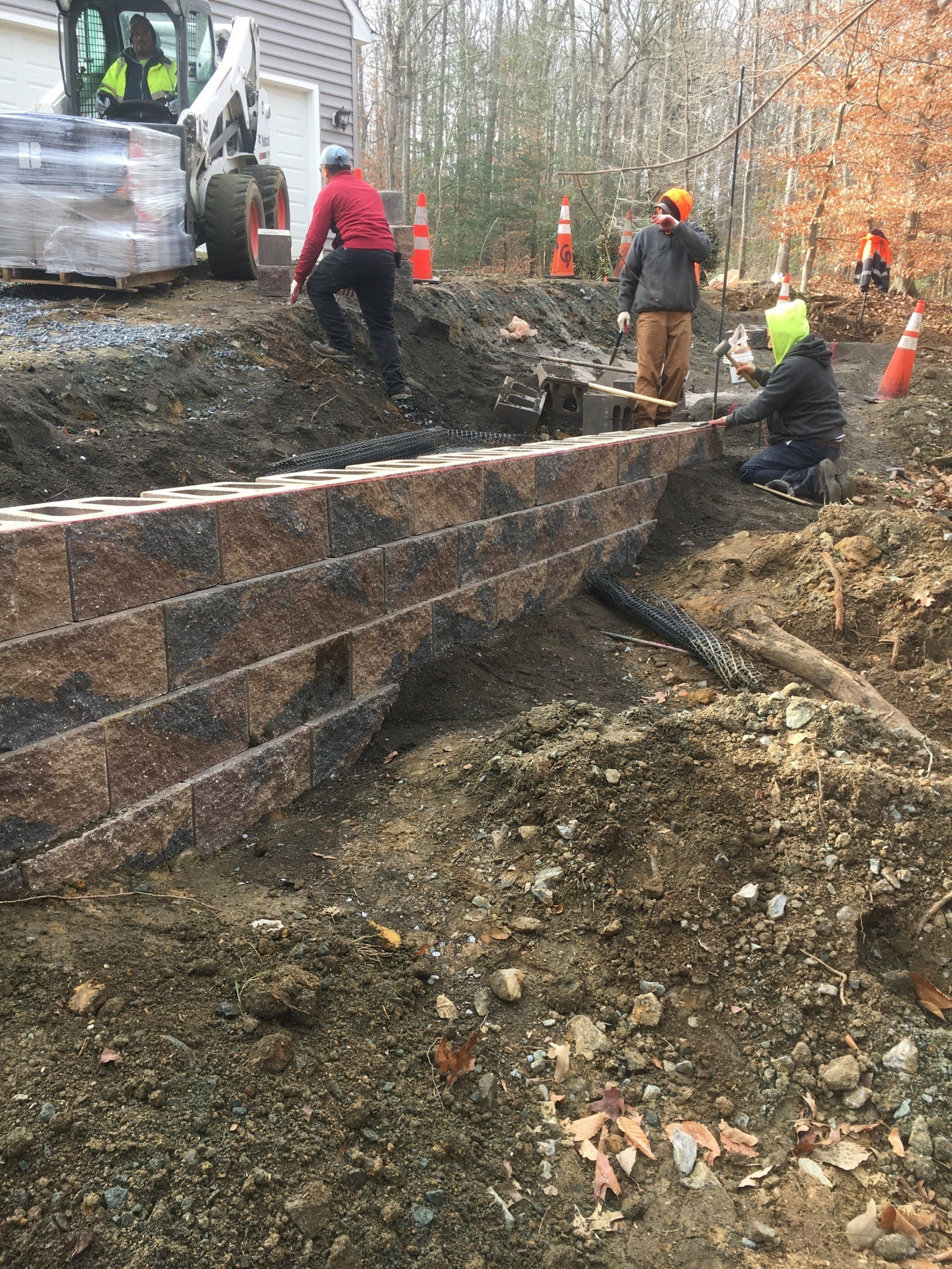 Construction workers building a retaining wall with blocks and a small excavator on a worksite.