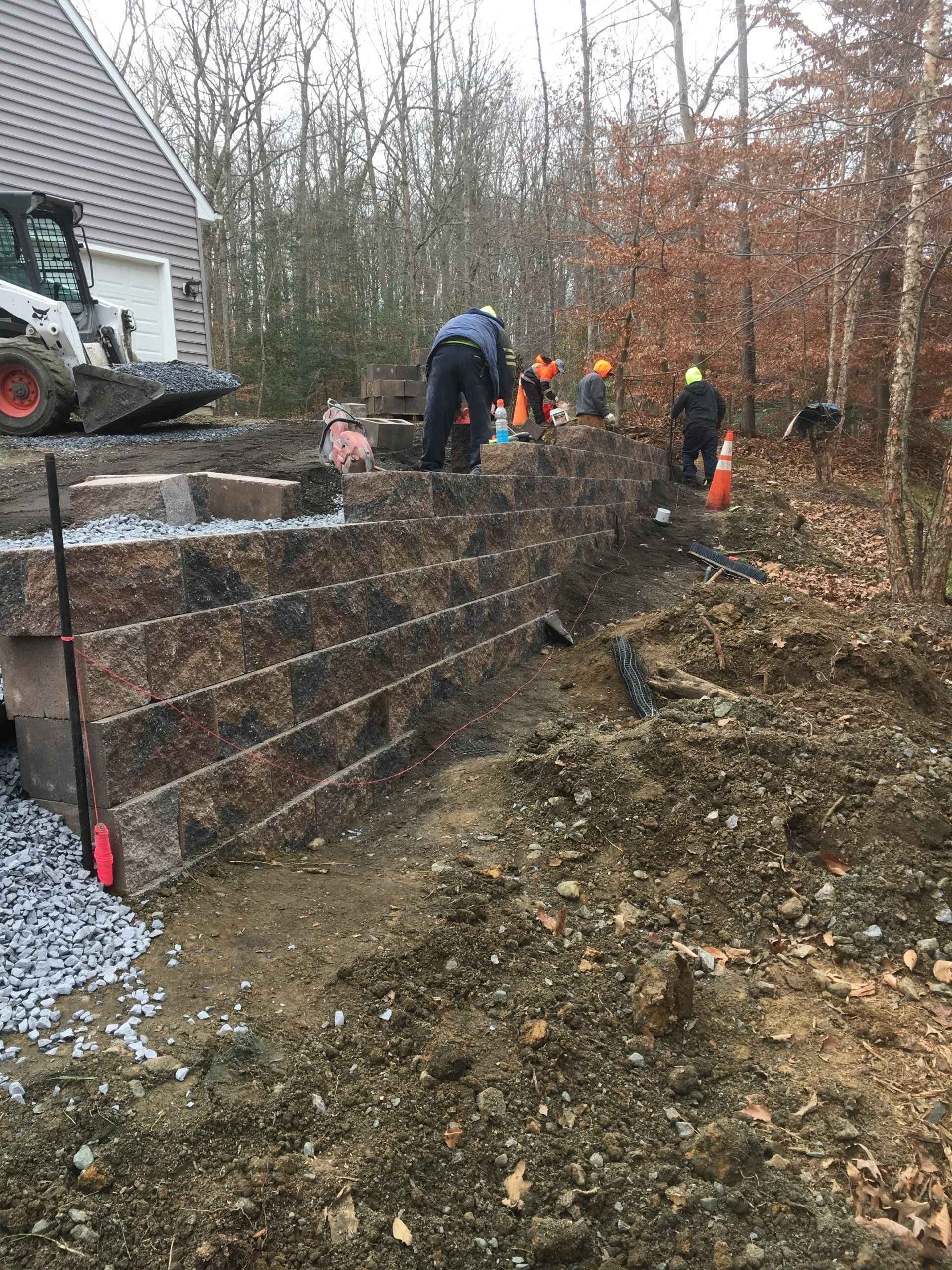 Construction workers building a retaining wall on a hillside. A skid steer sits nearby, and trees are in the background.