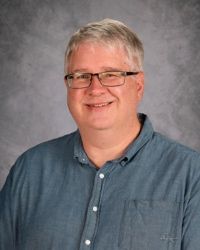 Smiling person in glasses and a blue button-up shirt against a gray studio backdrop