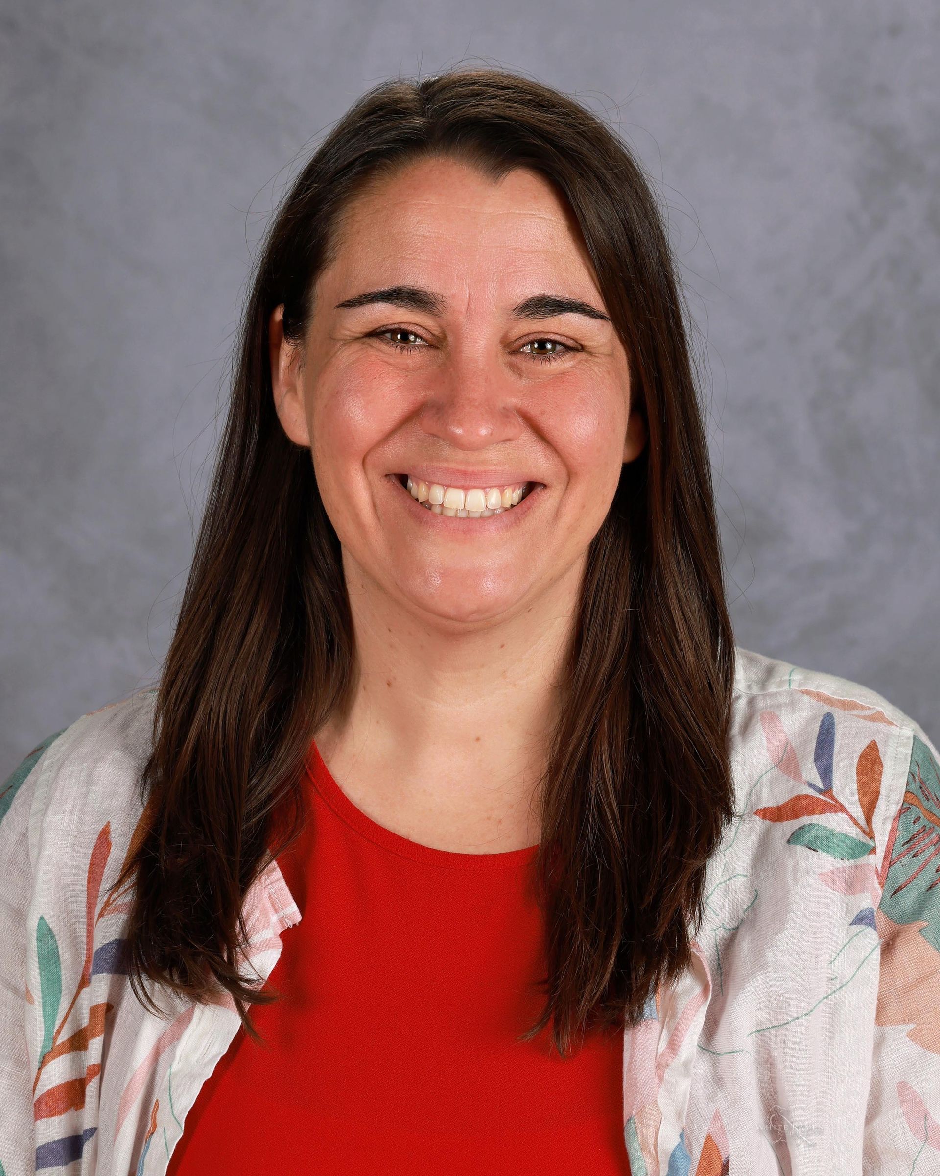 Woman with brown hair, smiling, wearing a red shirt and patterned cardigan.