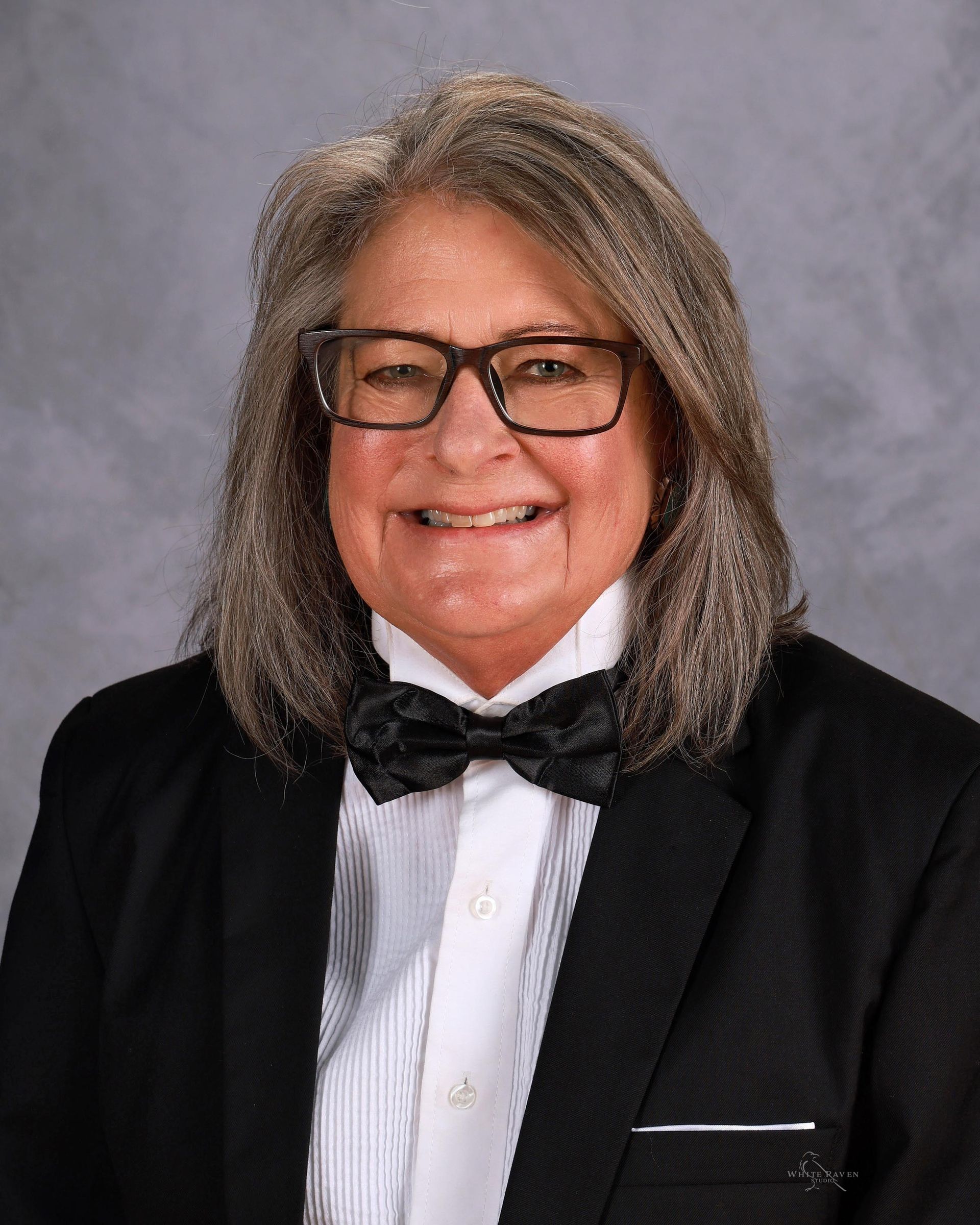 Person with long gray hair and glasses, wearing a tuxedo with a black bow tie, smiling.
