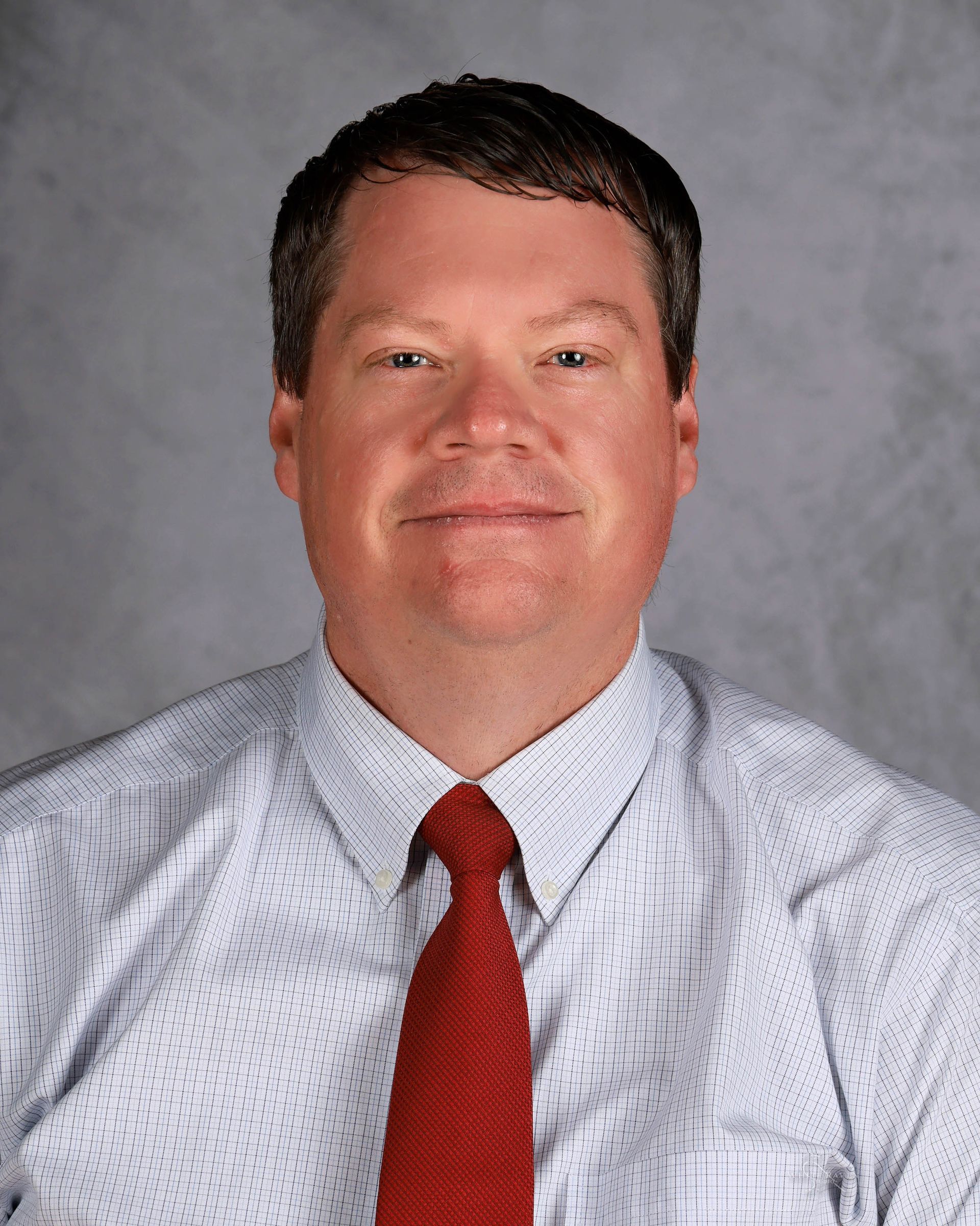 Man in light blue striped shirt and red tie, smiling, against a grey backdrop.