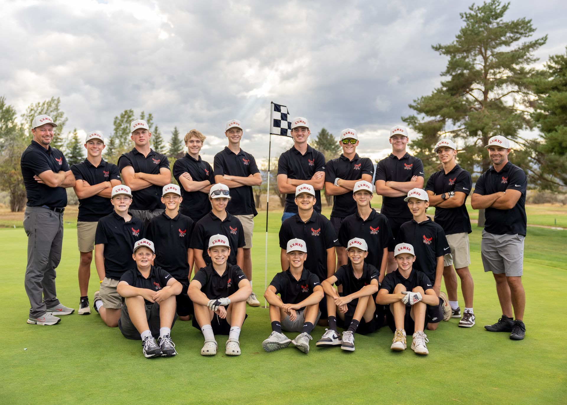 Golf team posing on a green course, wearing matching black shirts and caps, with trees and cloudy sky behind.