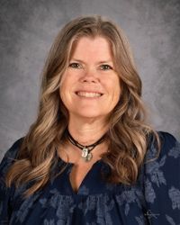 Smiling woman with long wavy hair against a gray studio backdrop, wearing a dark blue top and necklace