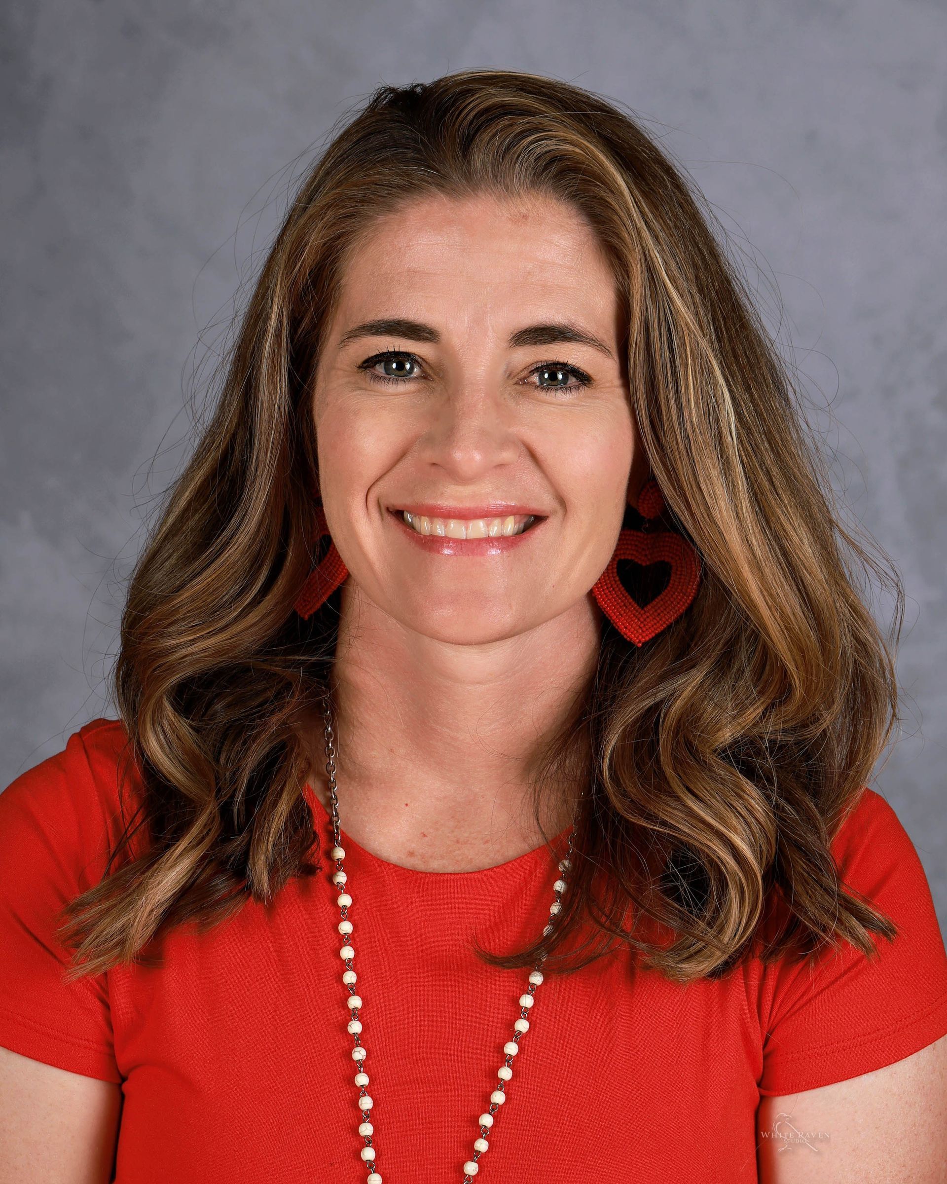 Woman with long brown hair, wearing a red shirt and earrings, smiling.