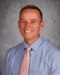 Smiling man in a blue shirt and pink tie against a gray studio background