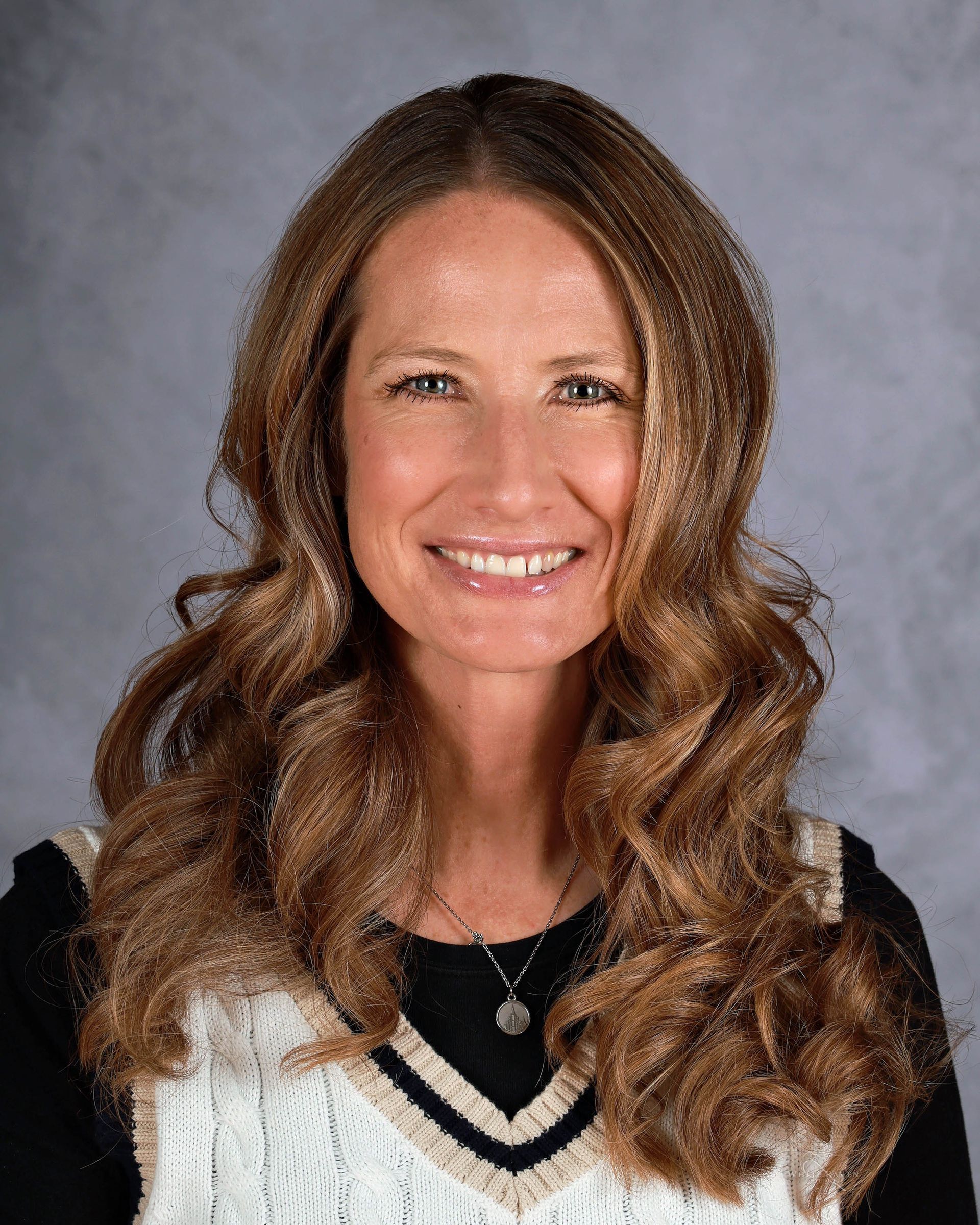Woman with long brown hair, smiling, wearing a black shirt and cream sweater vest, on a gray background.