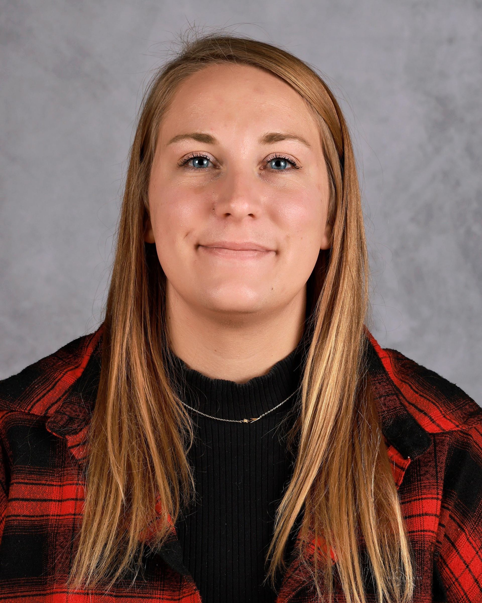 Blonde woman in red plaid shirt smiles, looking directly at the camera. Gray background.