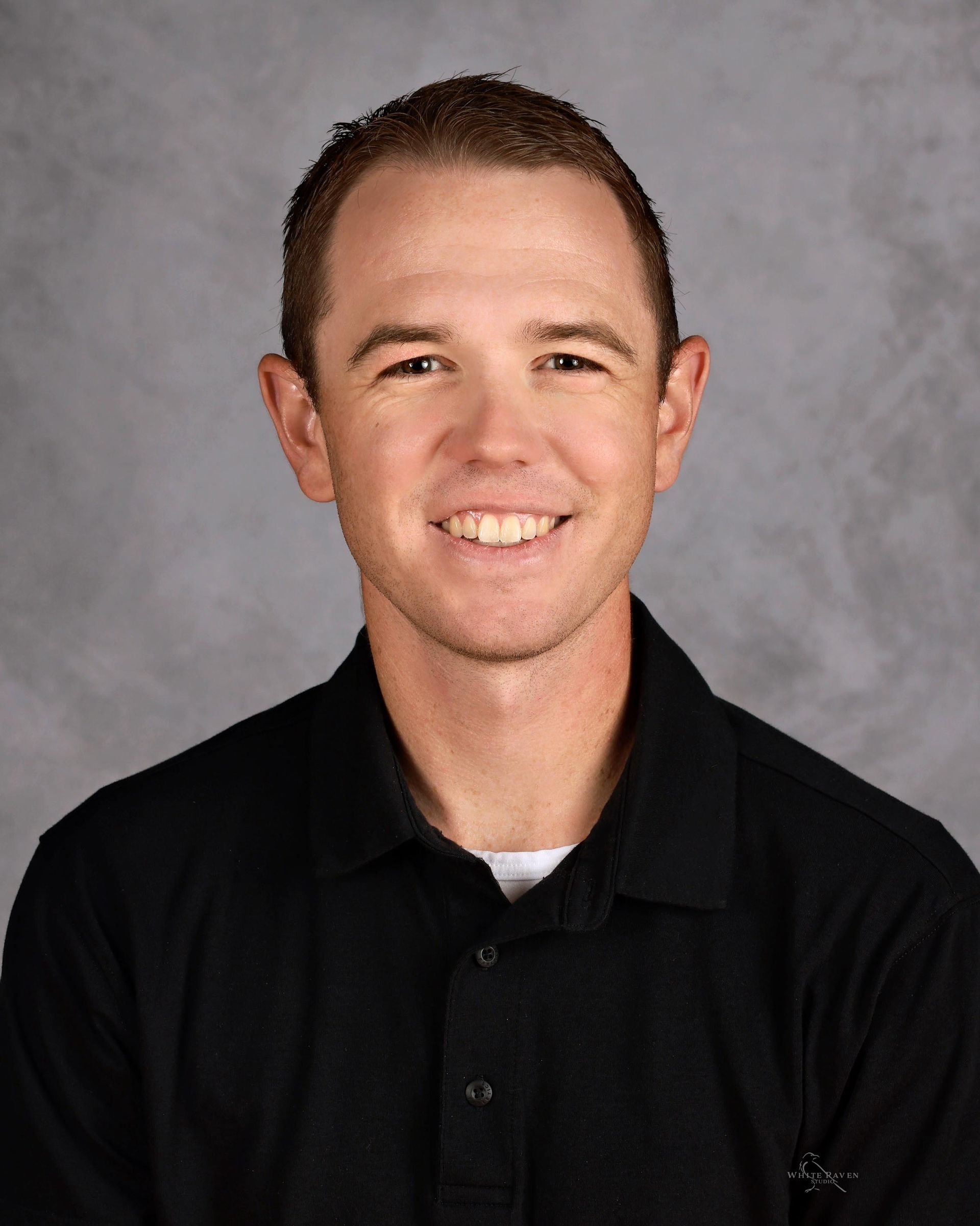 Man smiling, wearing a black collared shirt, against a gray background.