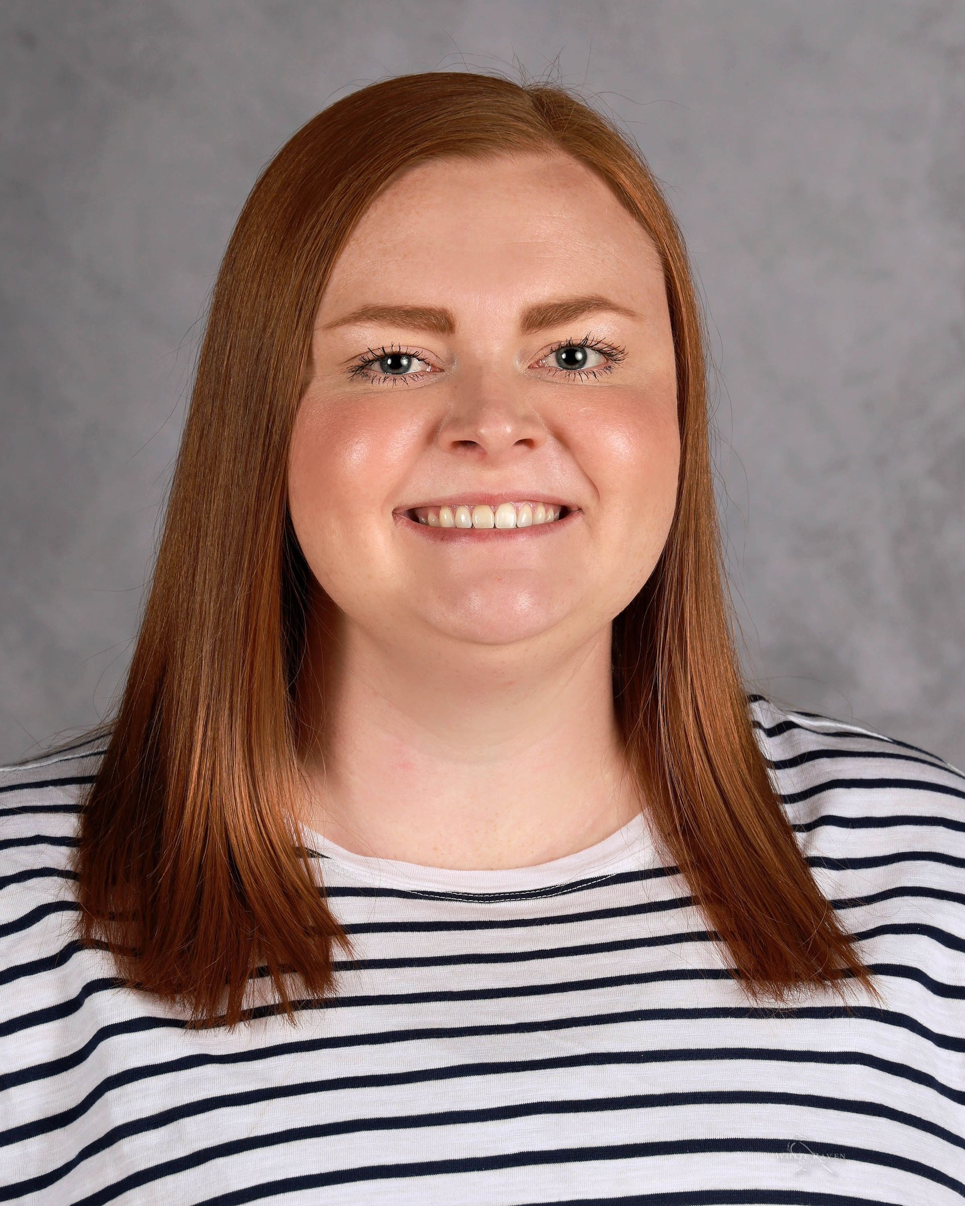 Woman with auburn hair smiles at the camera, wearing a striped shirt, against a gray backdrop.
