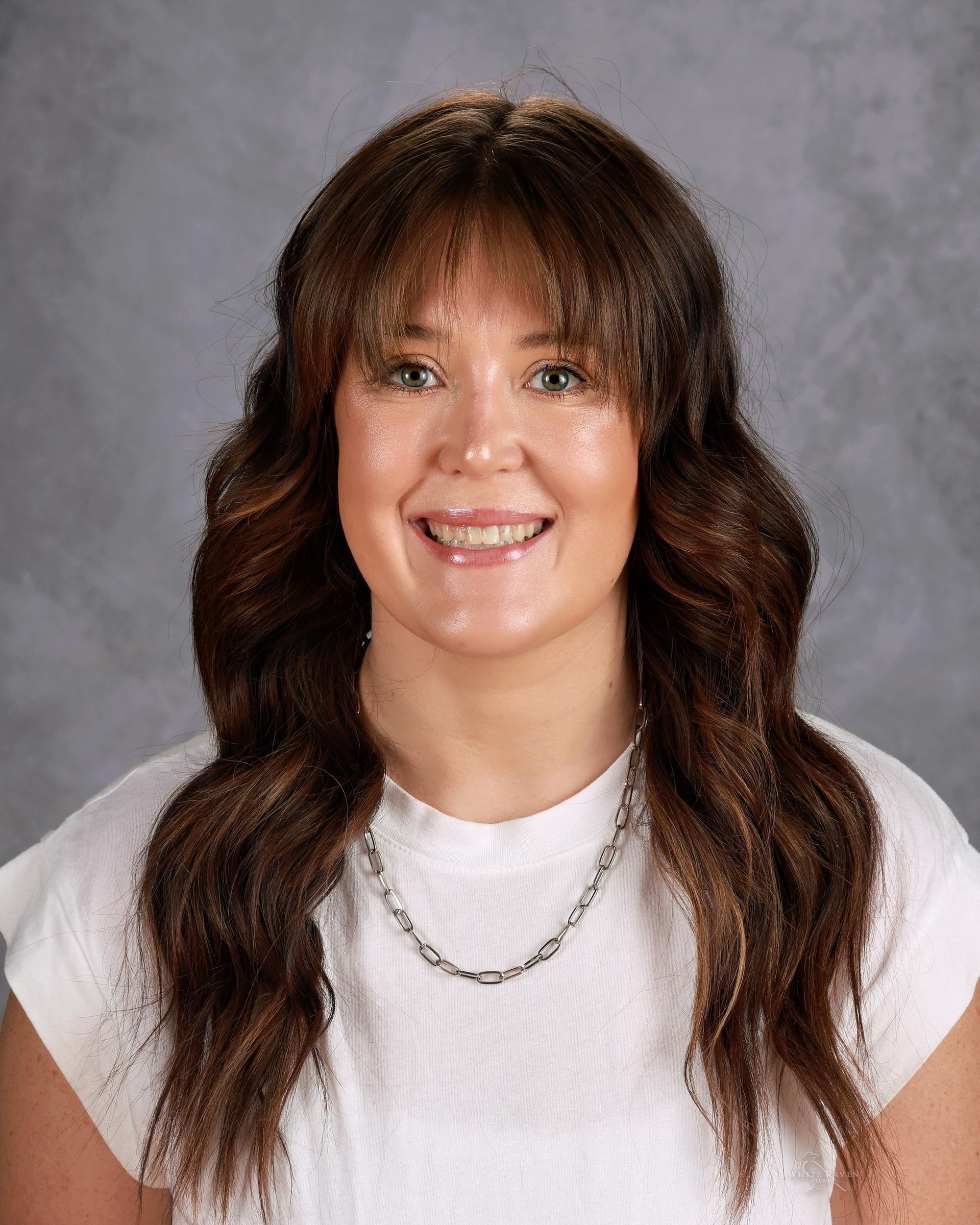 Woman with long brown hair and bangs smiles, wearing a white top and silver necklace.