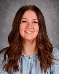 Smiling person with long brown hair wearing a light blue blouse against a gray backdrop