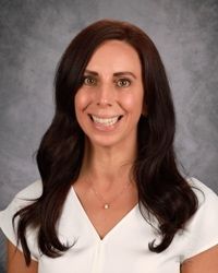 Portrait of a smiling person with dark wavy hair in a white top against a gray studio background