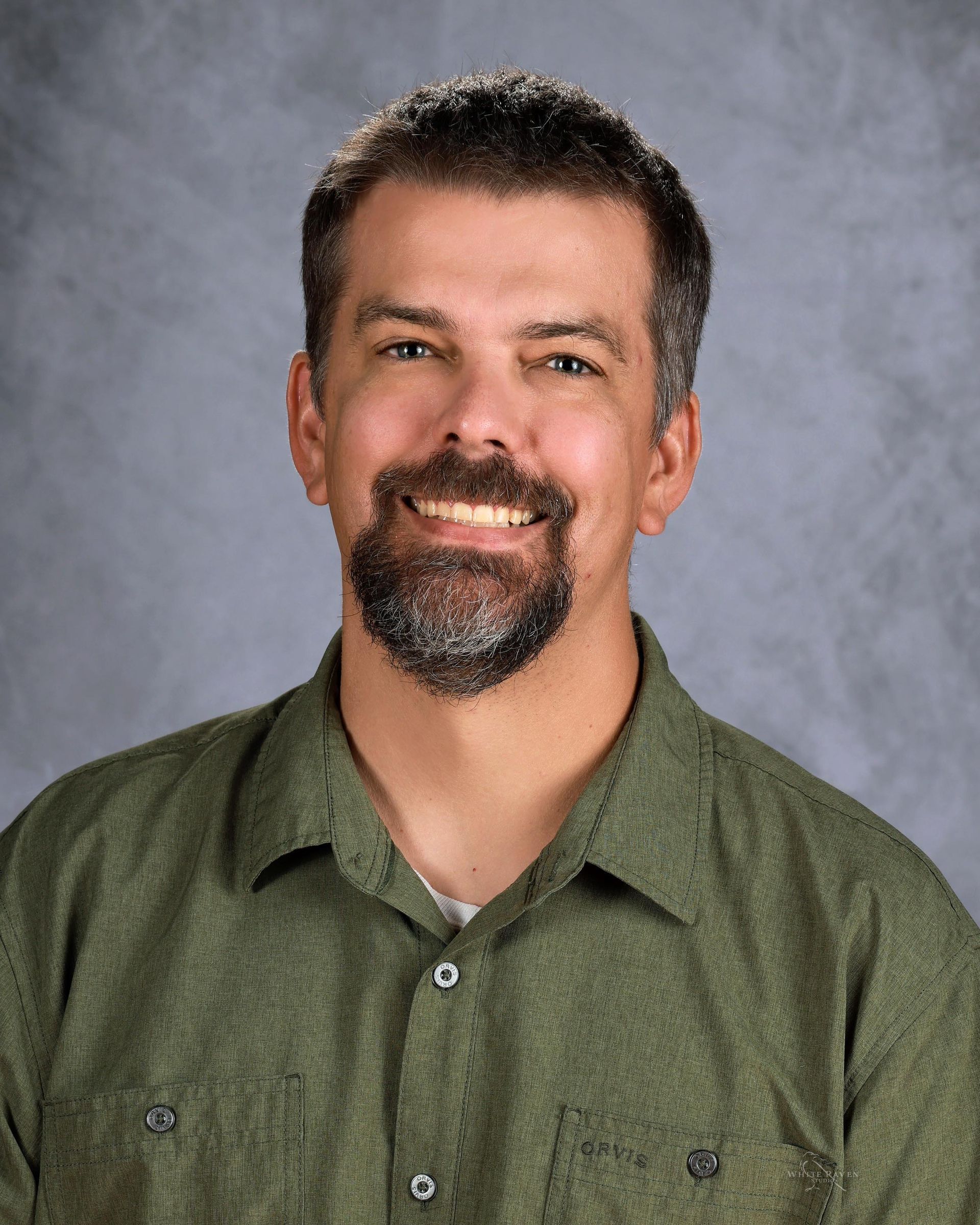 Smiling man with a salt-and-pepper beard and a green button-up shirt, against a gray background.