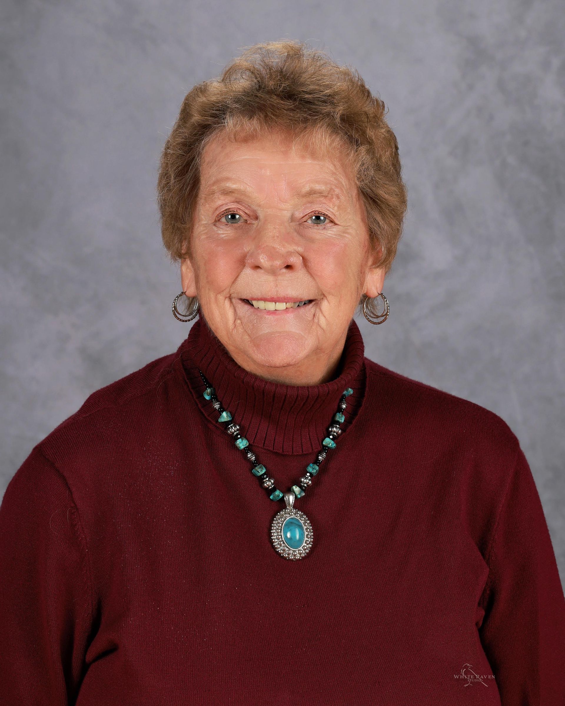 Smiling older woman with short brown hair wearing a maroon sweater and turquoise necklace.