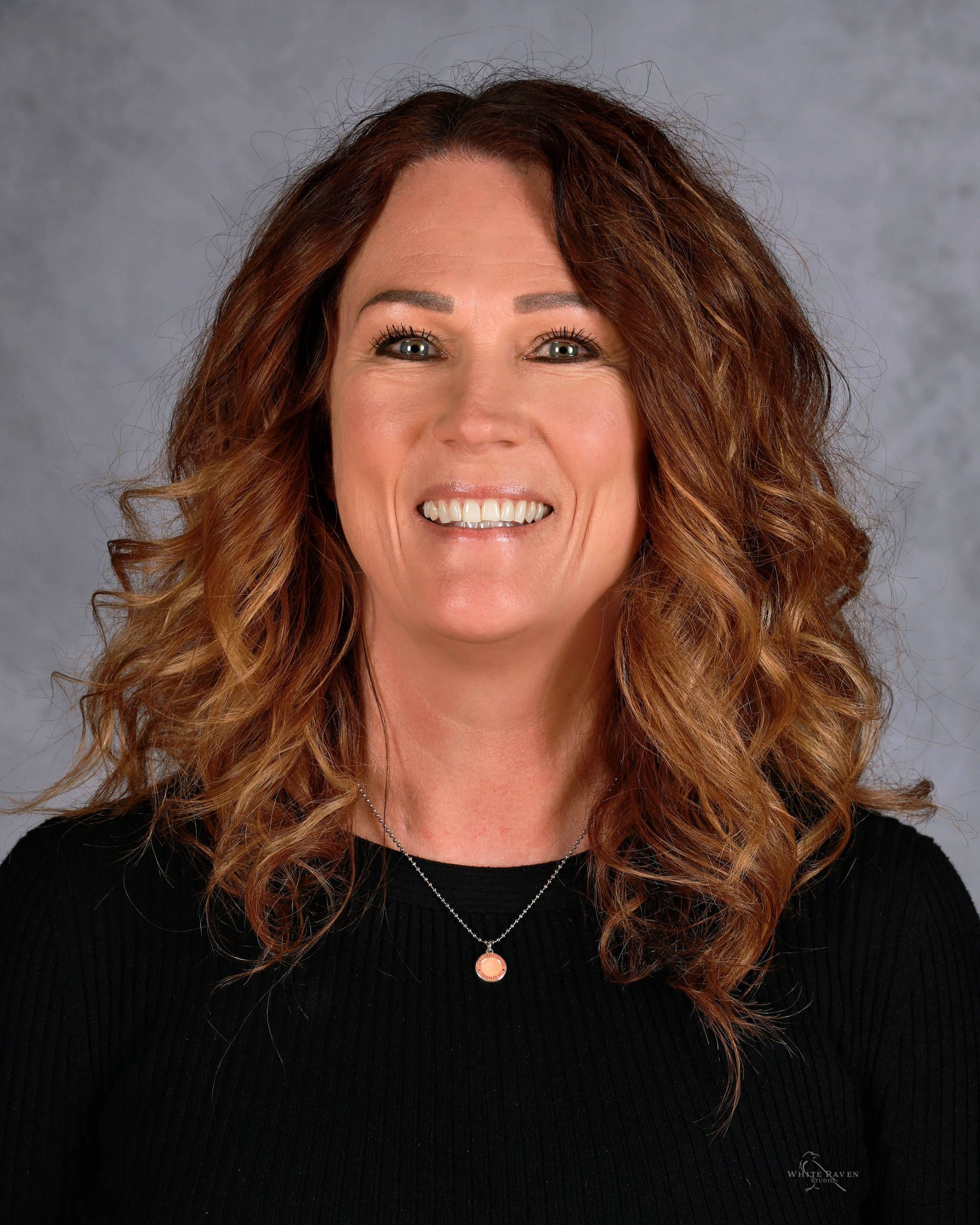 Woman with curly auburn hair smiles, wearing a black shirt and a necklace, in front of a gray background.
