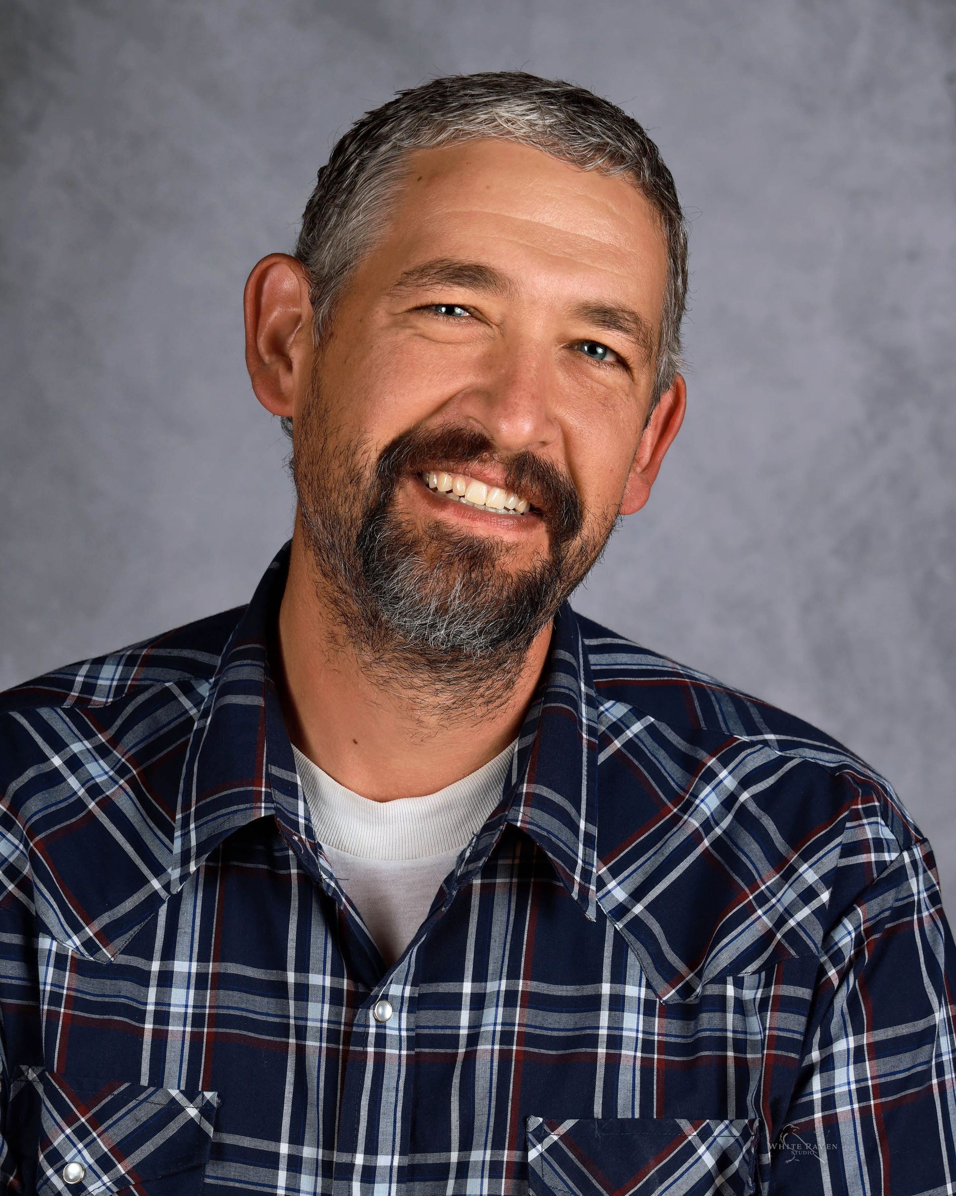 Smiling man with grey beard, wearing blue plaid shirt, posing in front of grey backdrop.