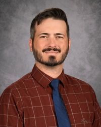 Portrait of a smiling man in a red plaid shirt and blue tie against a gray studio backdrop