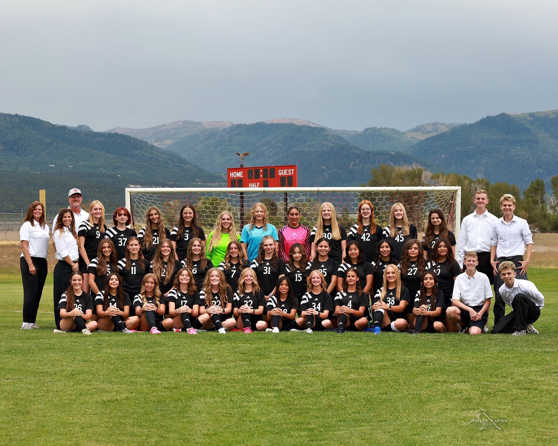 A soccer team poses in front of a goal on a grassy field with mountains in the background.