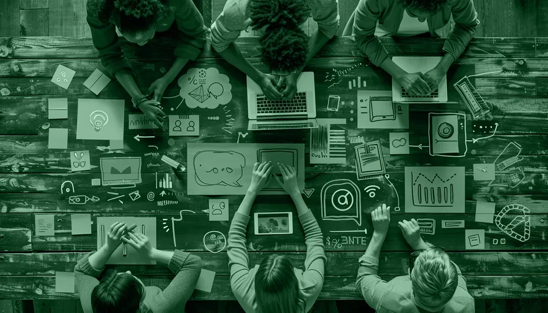 an overhead photo of people working on a large table computers and paper work are scattered all over the table top