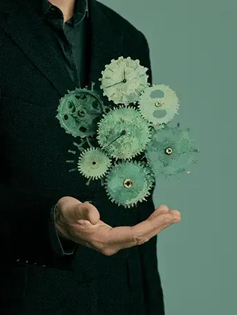 An extended hand from a figure in a dark green suit. Above his hand we see floating green gears in motion. The background is a muted and empty.