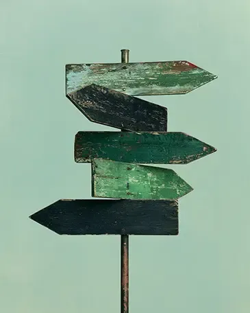 Woodened green directional signpost with five arrow-shaped boards on a pole pointing in various dis-coherent directions.