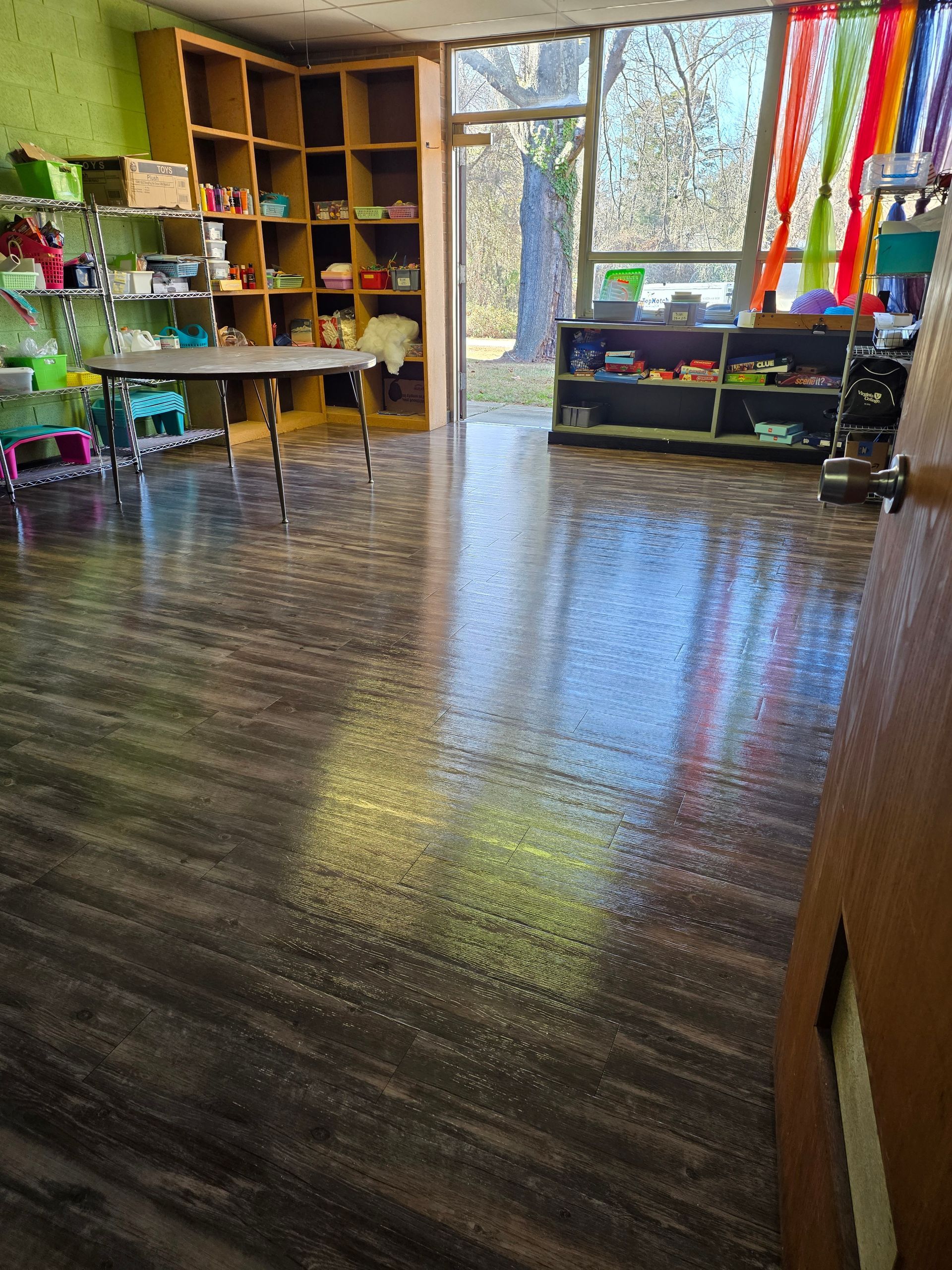 Empty classroom with a shiny wood floor, cubbies on the left, colorful ribbons, and bright windows at the back