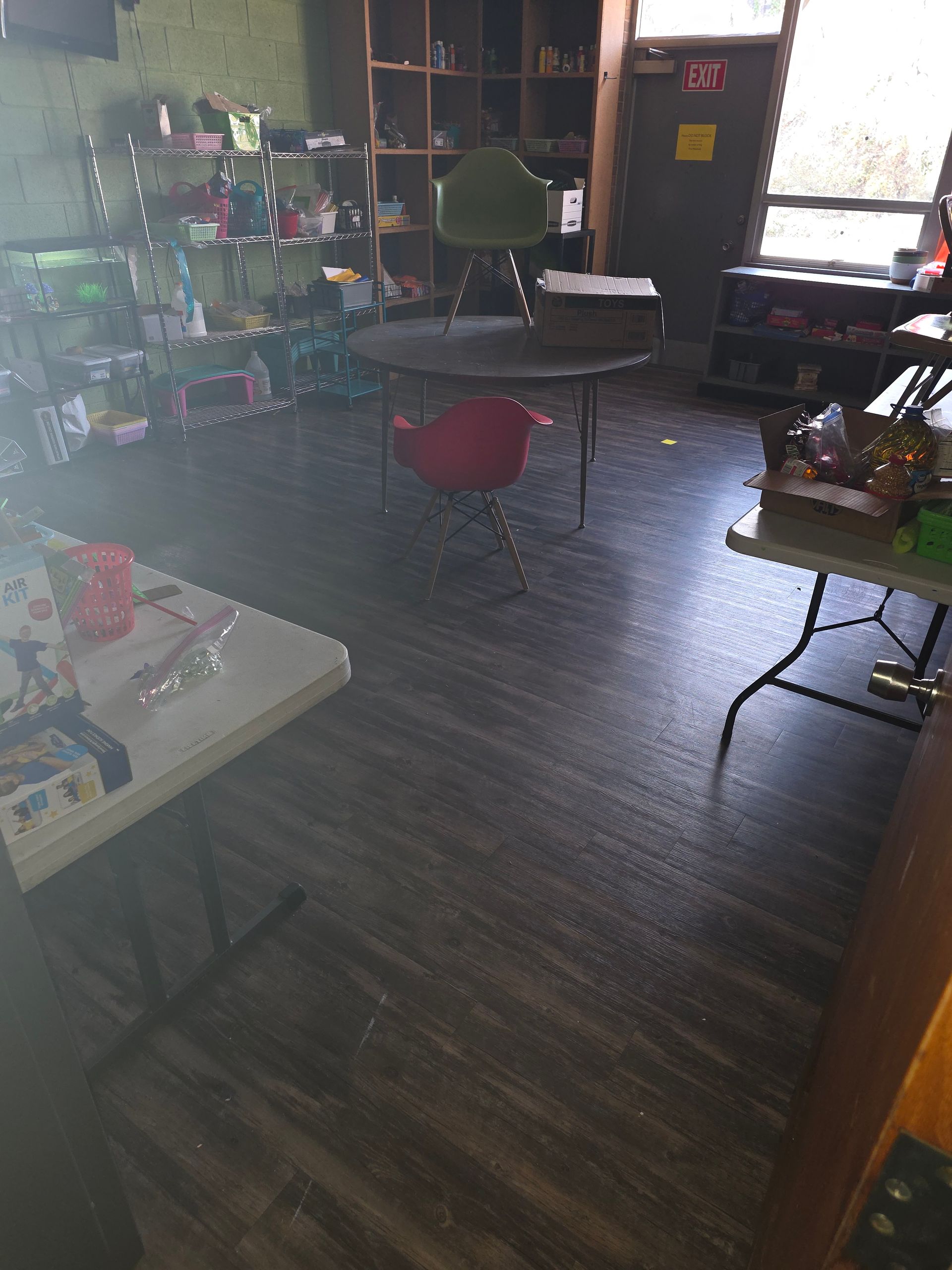 Dim classroom with tables, chairs, shelves, and sunlight from a window.